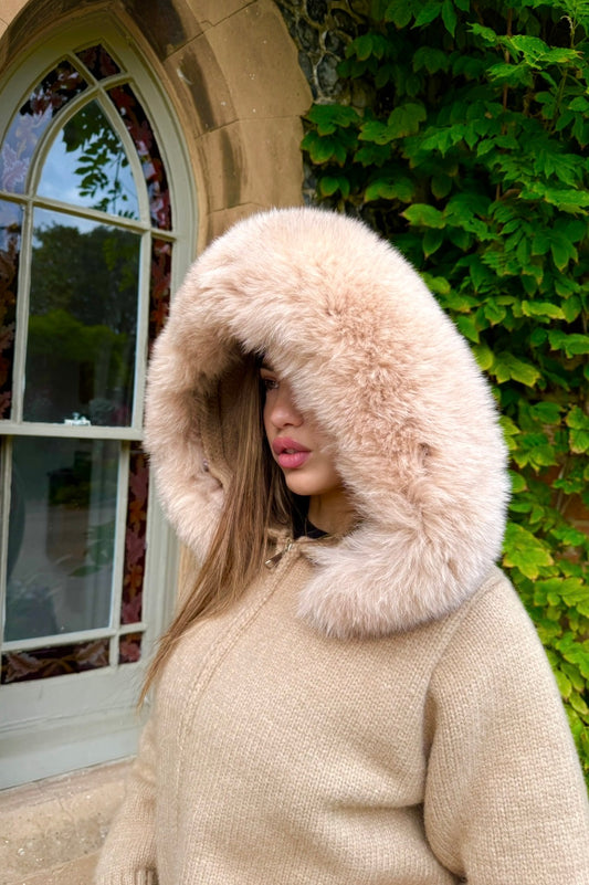 Person wearing a beige coat with a fur hood in front of a building with arched windows.