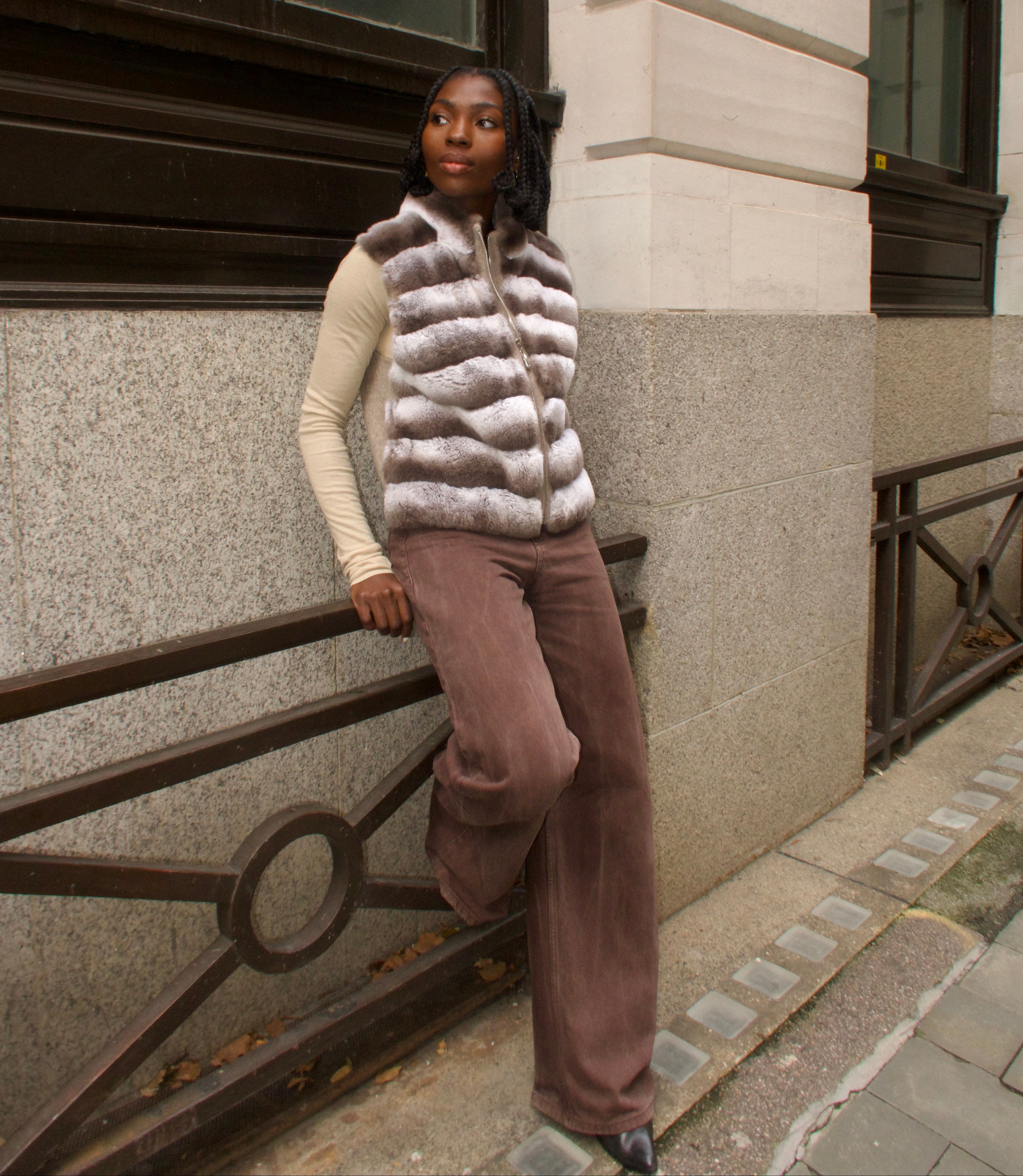 Woman wearing a fur vest and brown pants standing against a building.