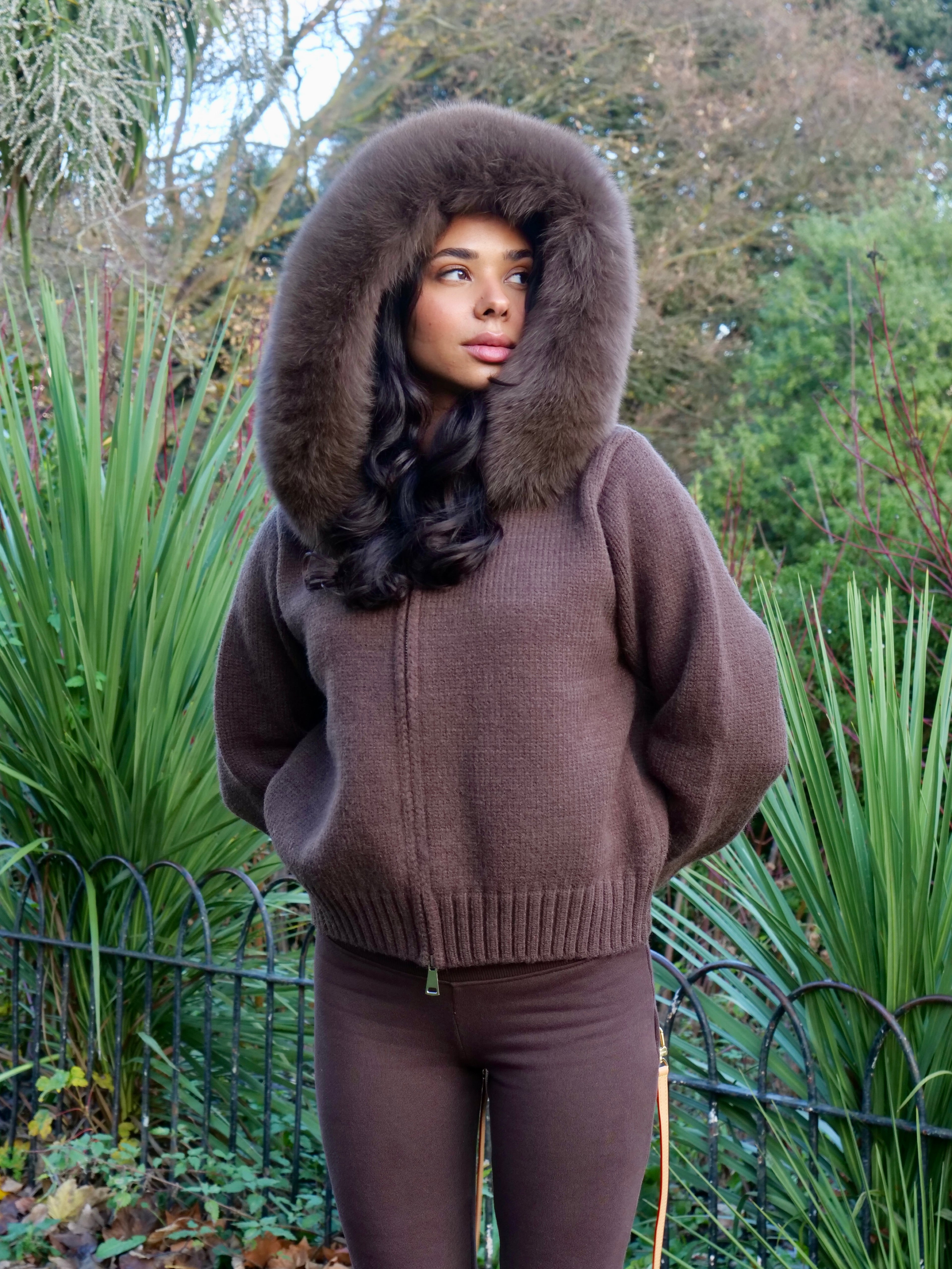 Person wearing a brown knitted outfit with a fur-lined hood in an outdoor setting.