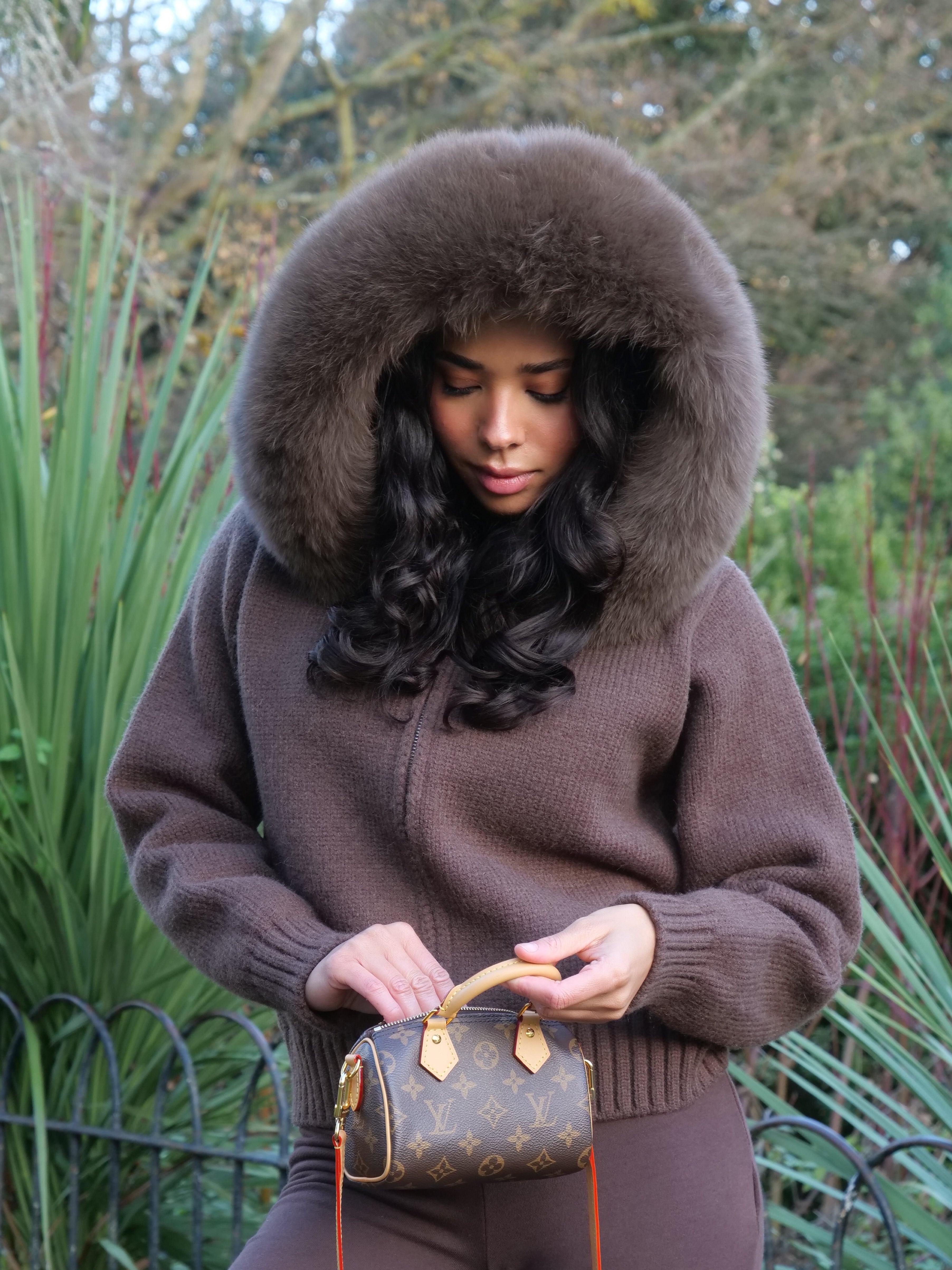 Woman in brown outfit with fur hood holding a vintage-style handbag in a garden setting
