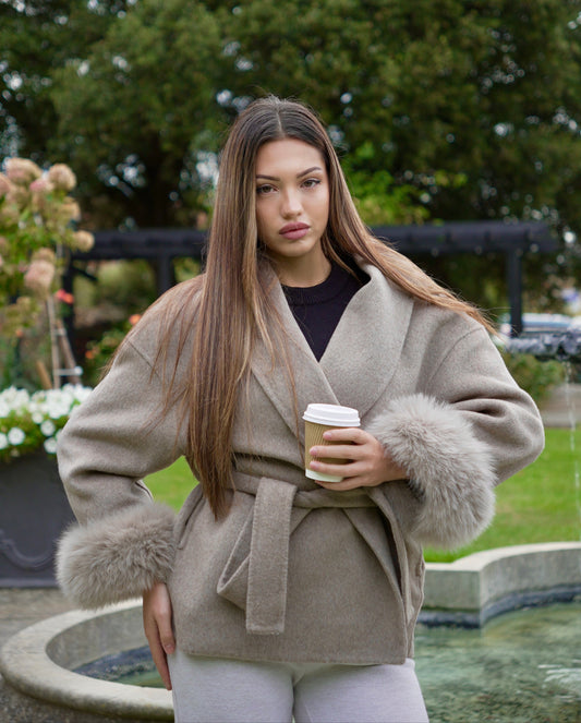 Woman in a beige coat with fur trim holding a coffee cup outdoors.