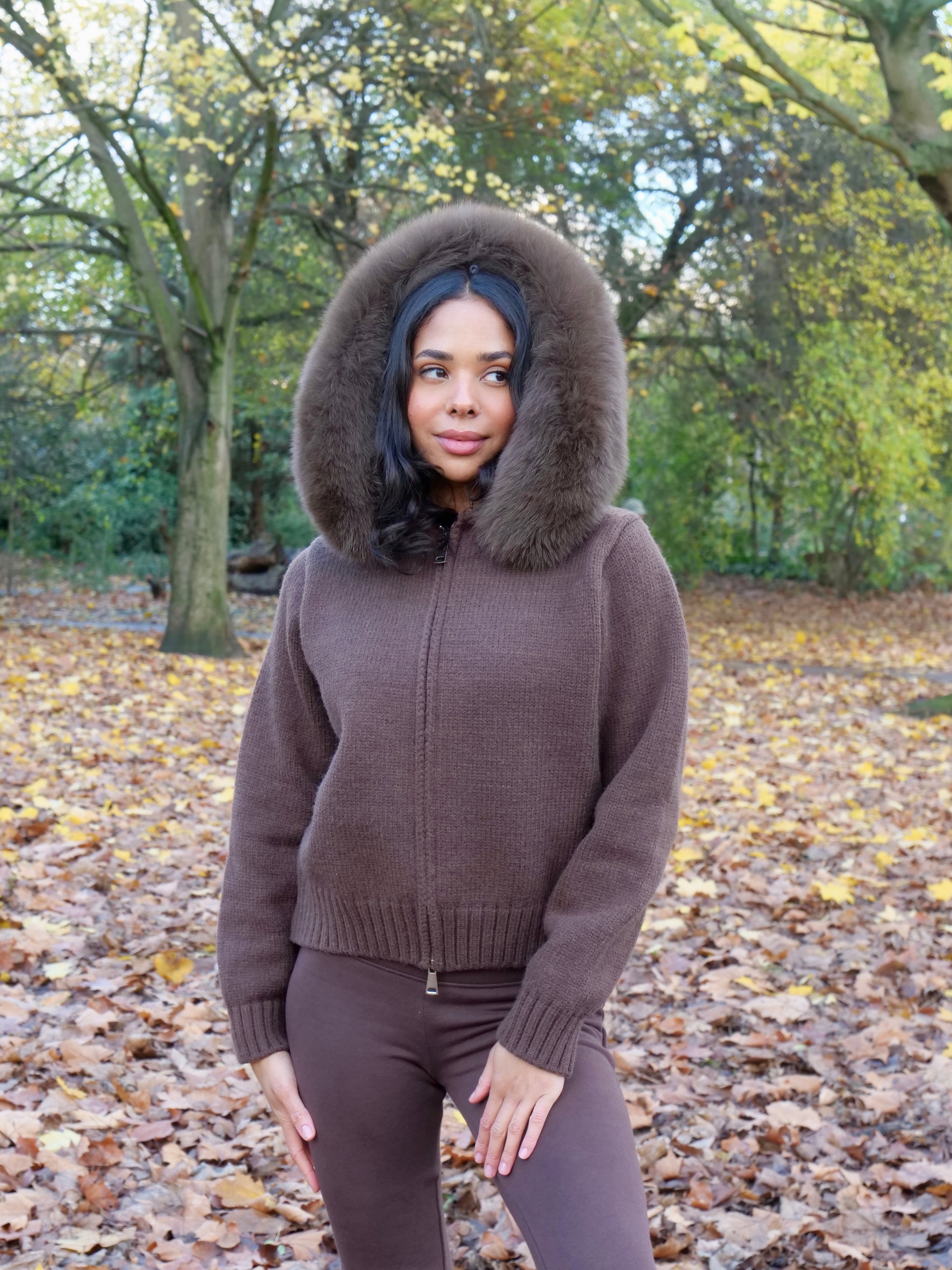 Woman wearing a brown hoodie with a fox fur collar and pants in a park with trees and fallen leaves.