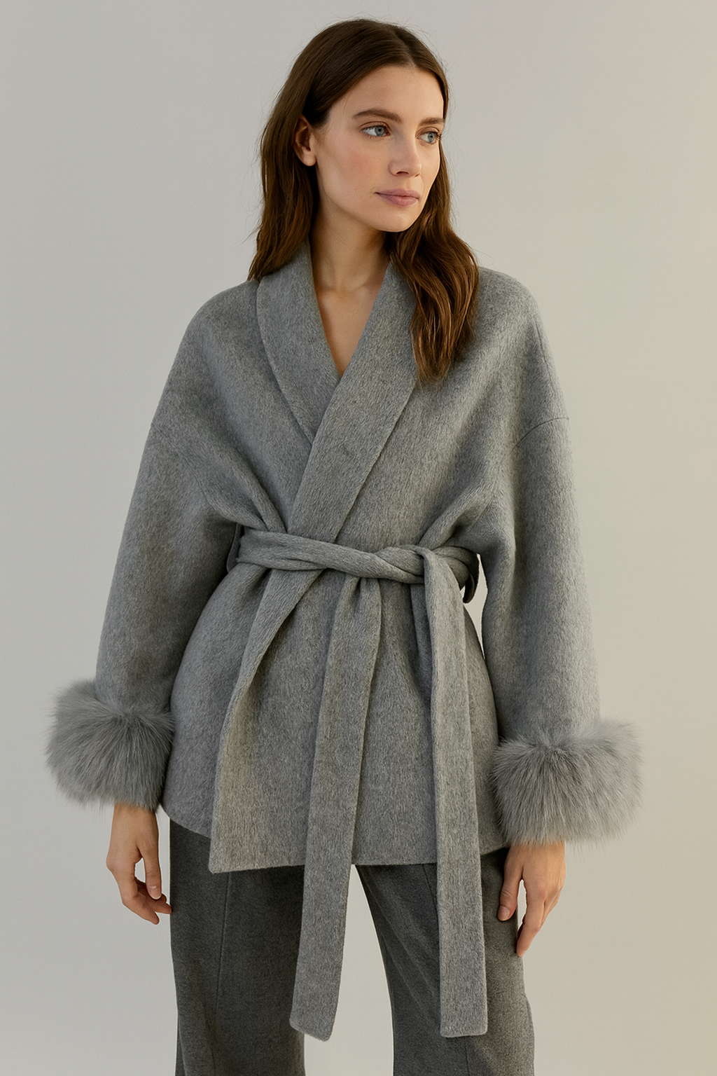 Woman wearing a gray wrap coat with fur cuffs on a plain background