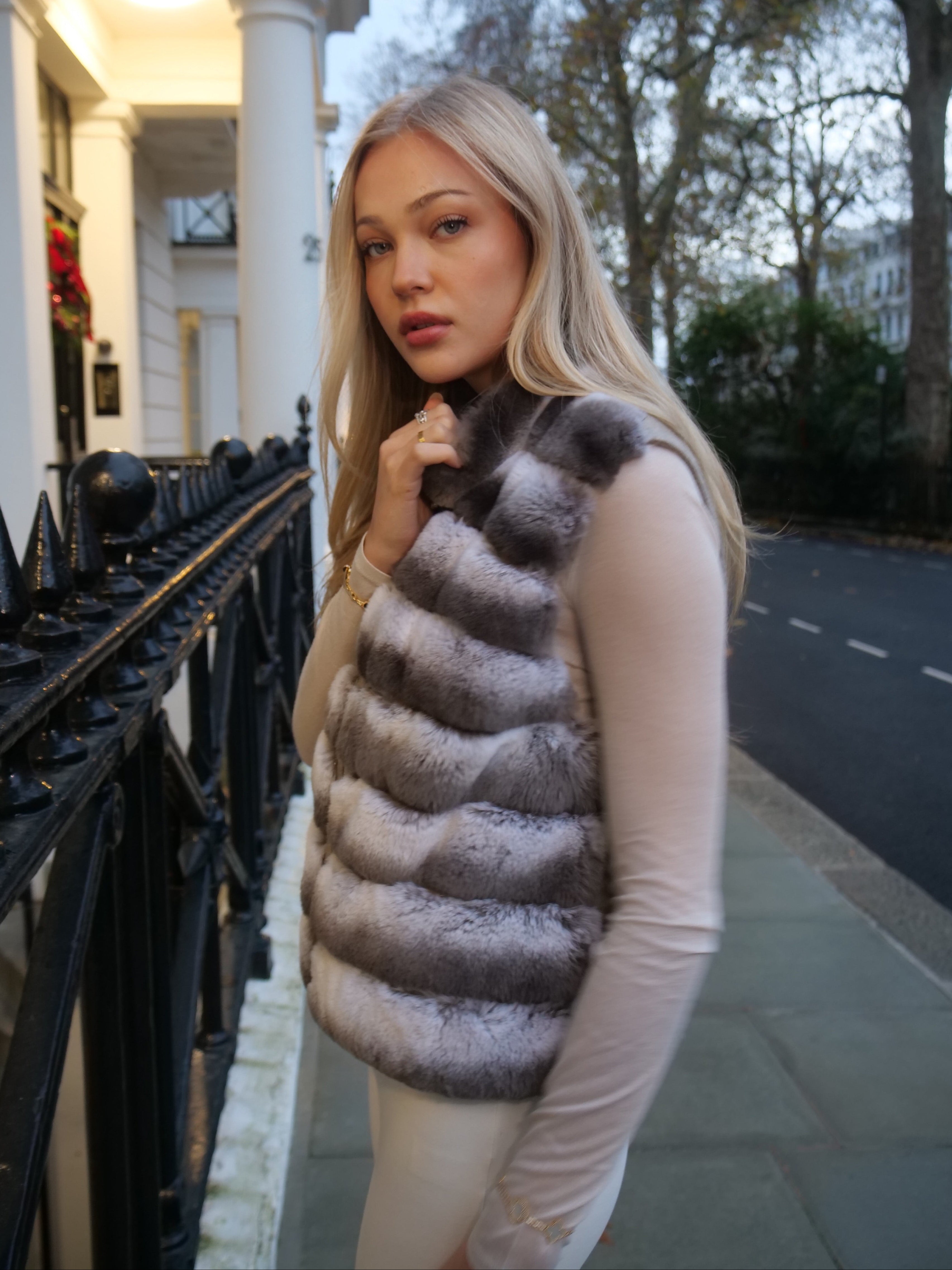 Woman wearing a fur vest standing on a street with buildings and trees in the background